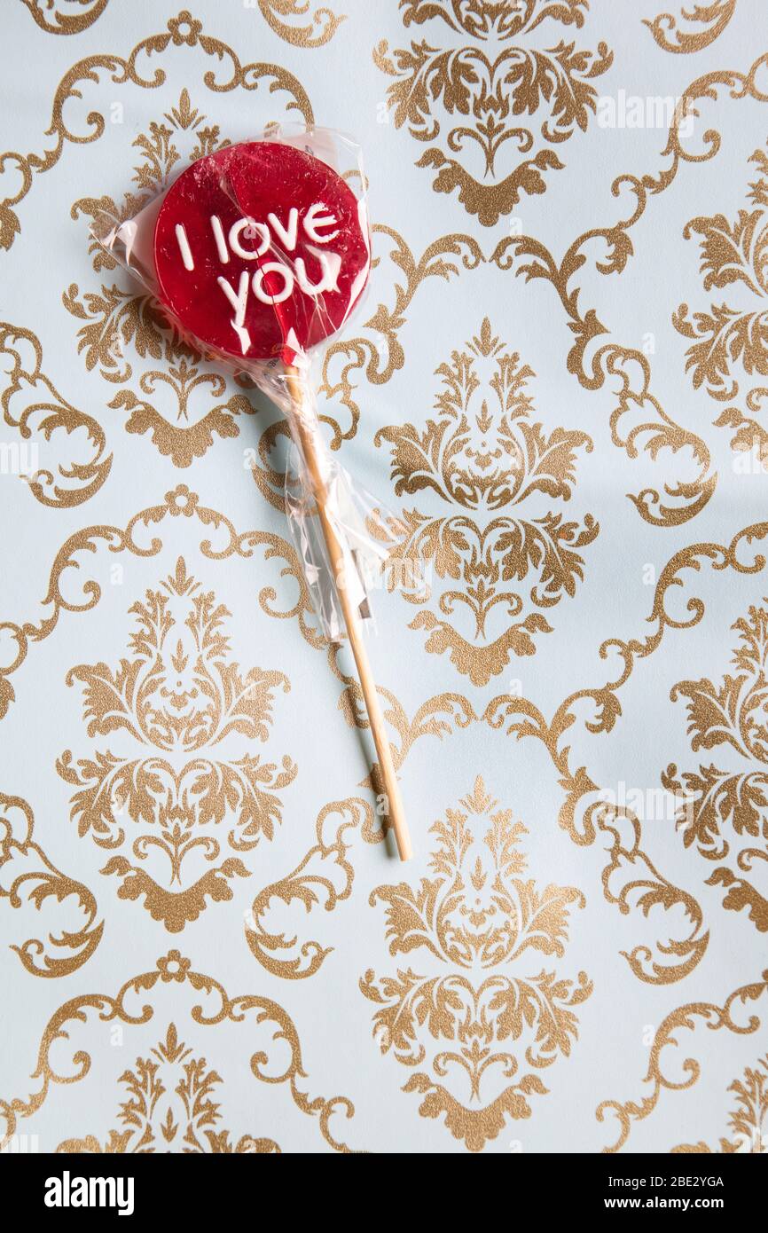 a sweet wrapped 'I Love you' red lollypop against a gold and light blue decorated background, flat lay, top view, minimalistic Stock Photo