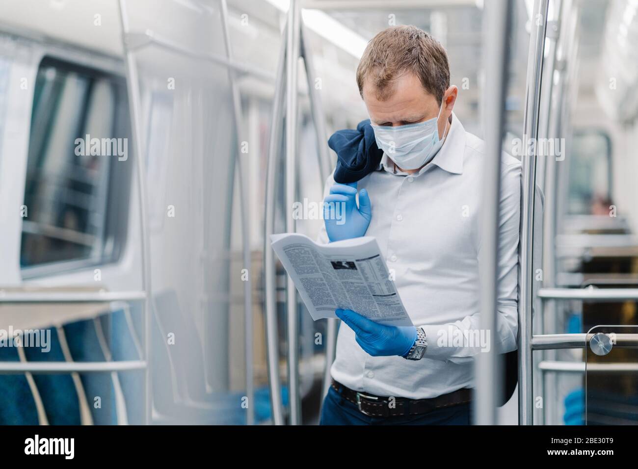 Coronavirus pandemic, Covid-19, quarantine measure. Serious adult man in white shirt, holds black jacket, concentrated in newspaper, wears protective Stock Photo