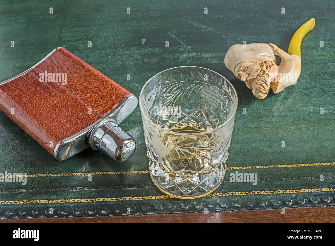 Hip flask, whisky glass, Meerschaum pipe on green leather table. Concept. Gentleman’s study, relaxation, social isolation, nostalgia, own company. Stock Photo