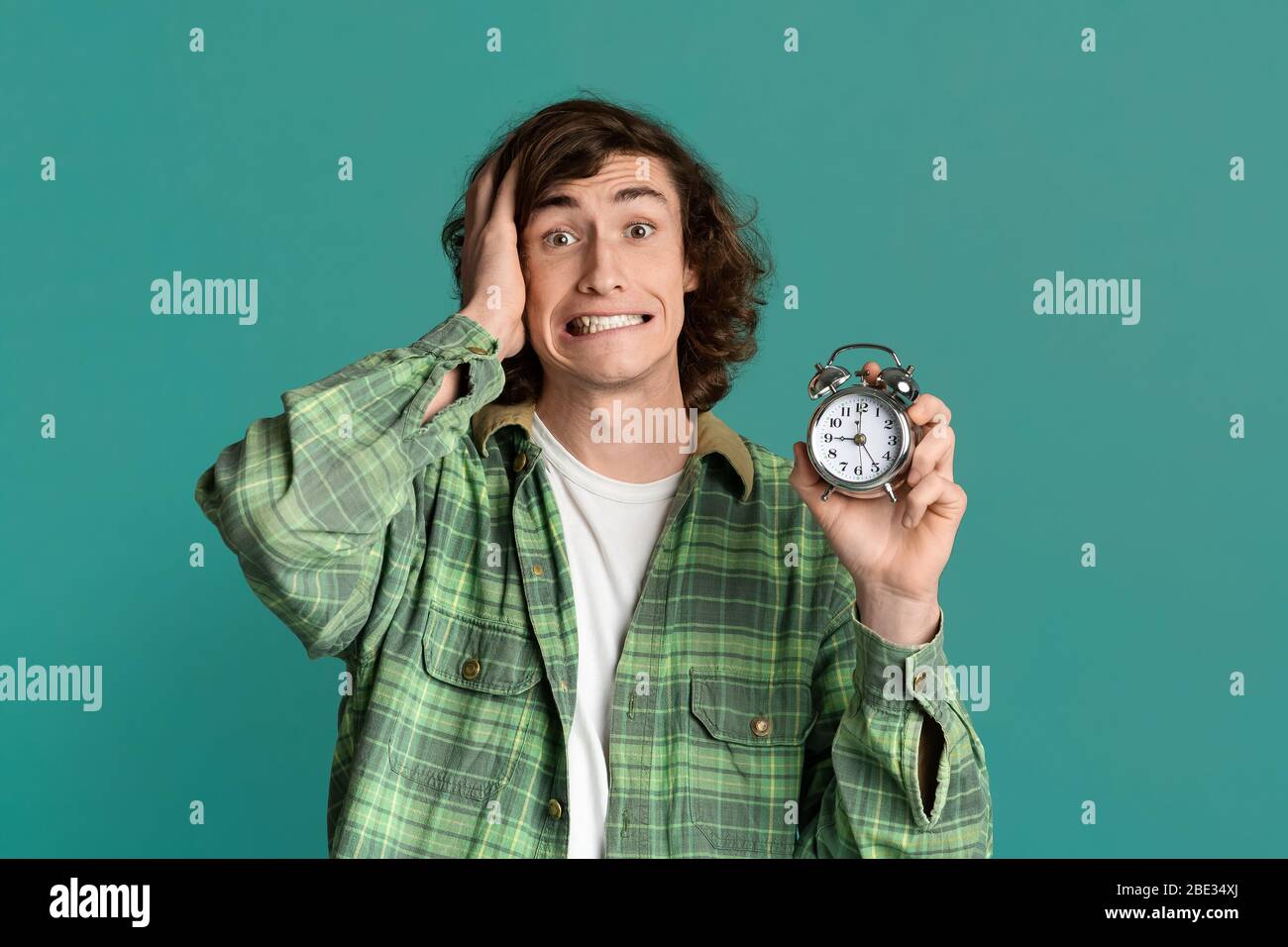 Time optimization. Young guy with alarm clock scared of being late on turquoise background Stock