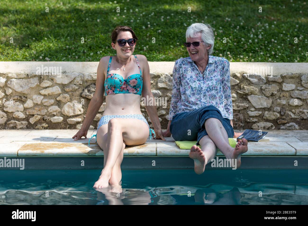 A mother and daughter enjoy sit on the side of a swimming pool in the sunshine. Stock Photo