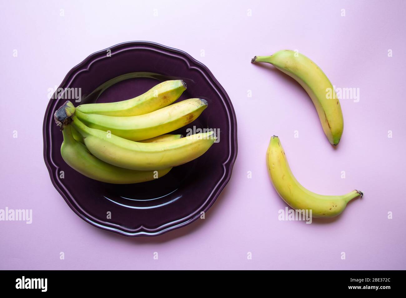 a stylish pastel coloured image of a bunch of bananas an a decorative purple plate with pink background, flat lay, top view Stock Photo