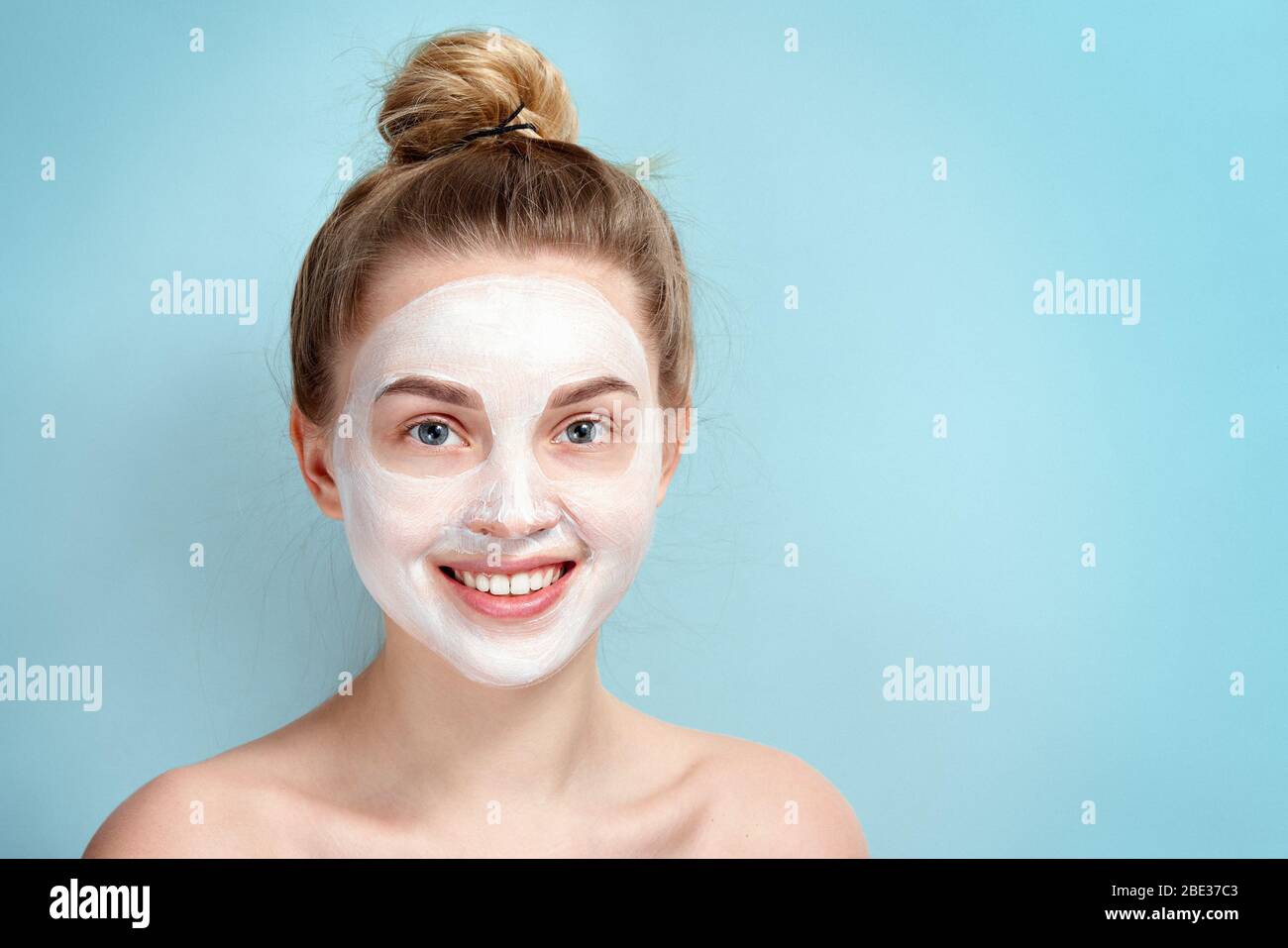 Young girl in a cosmetic mask of white smiling face. Skincare. Blonde woman smiling on blue background. moisturizer mask. Beautiful face of young adul Stock Photo