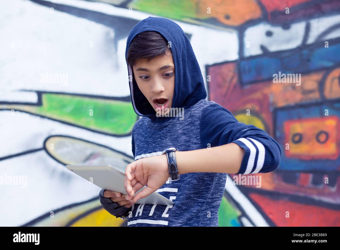 Boy, man in blue hoody holding tablet pad running late to school looking to his wrist watch isolated graffiti wall background. Outdoors, lifestyle hor Stock Photo