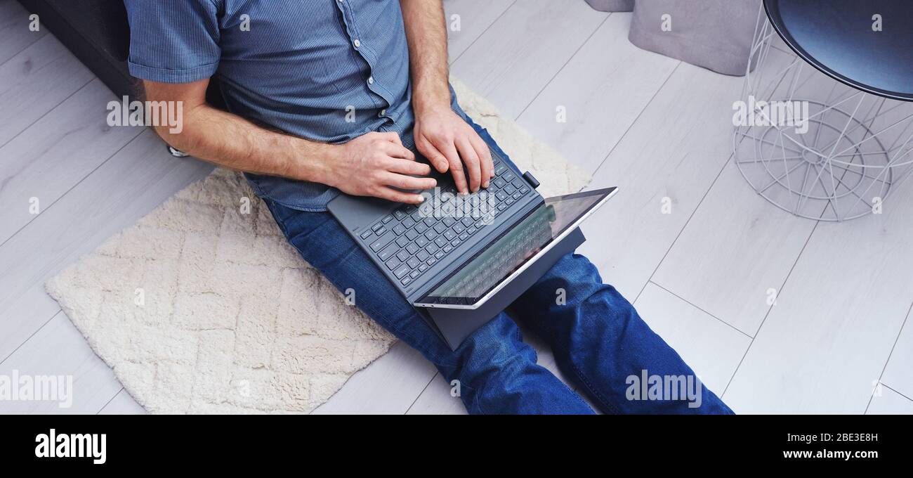 Young man in casual clothes sitting on floor and working on his laptop computer at home. Stay home and work online. Quarantine concept, panoramic Stock Photo