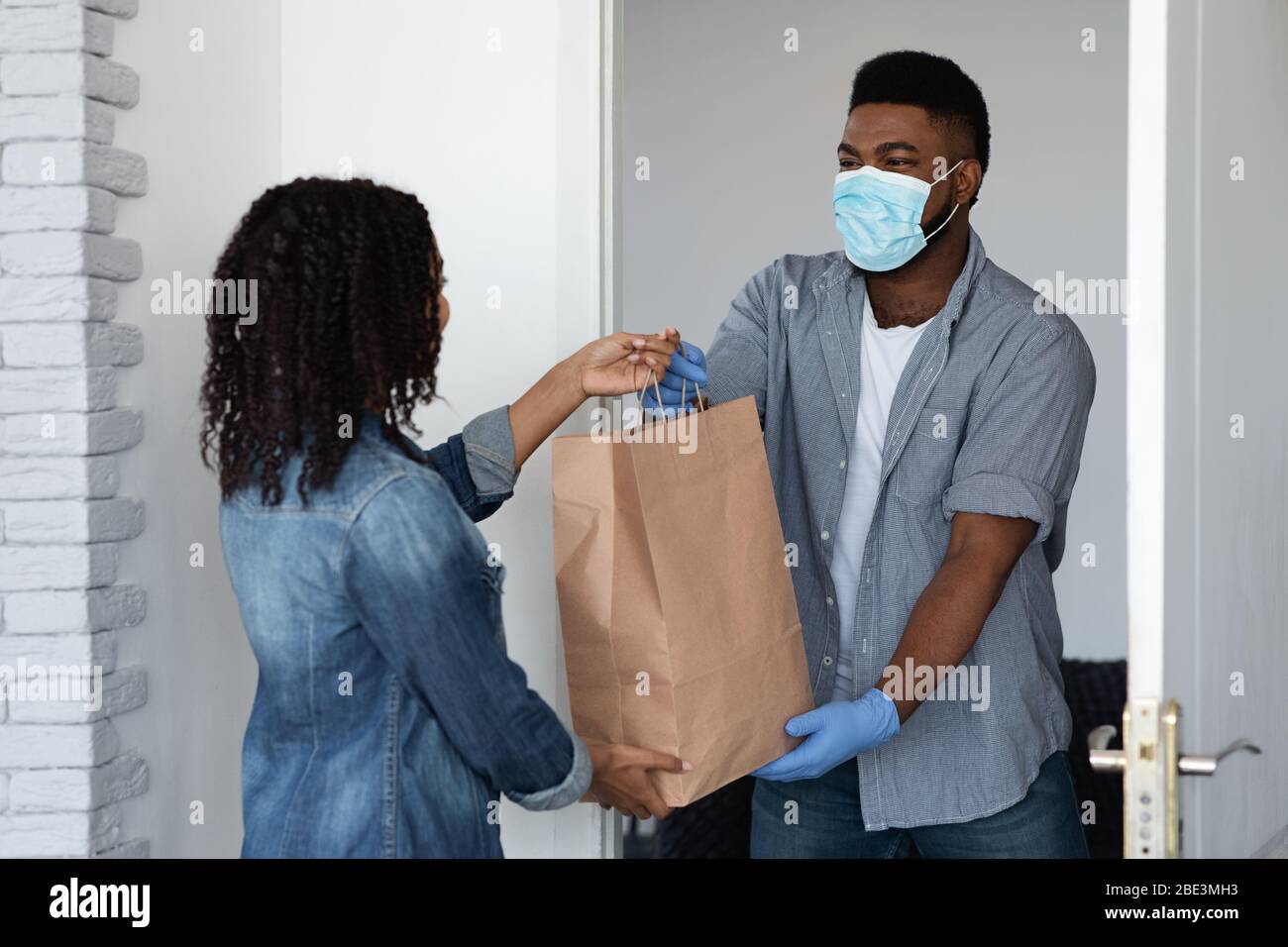 Black courier guy in medical mask delivering grocery order to woman Stock Photo