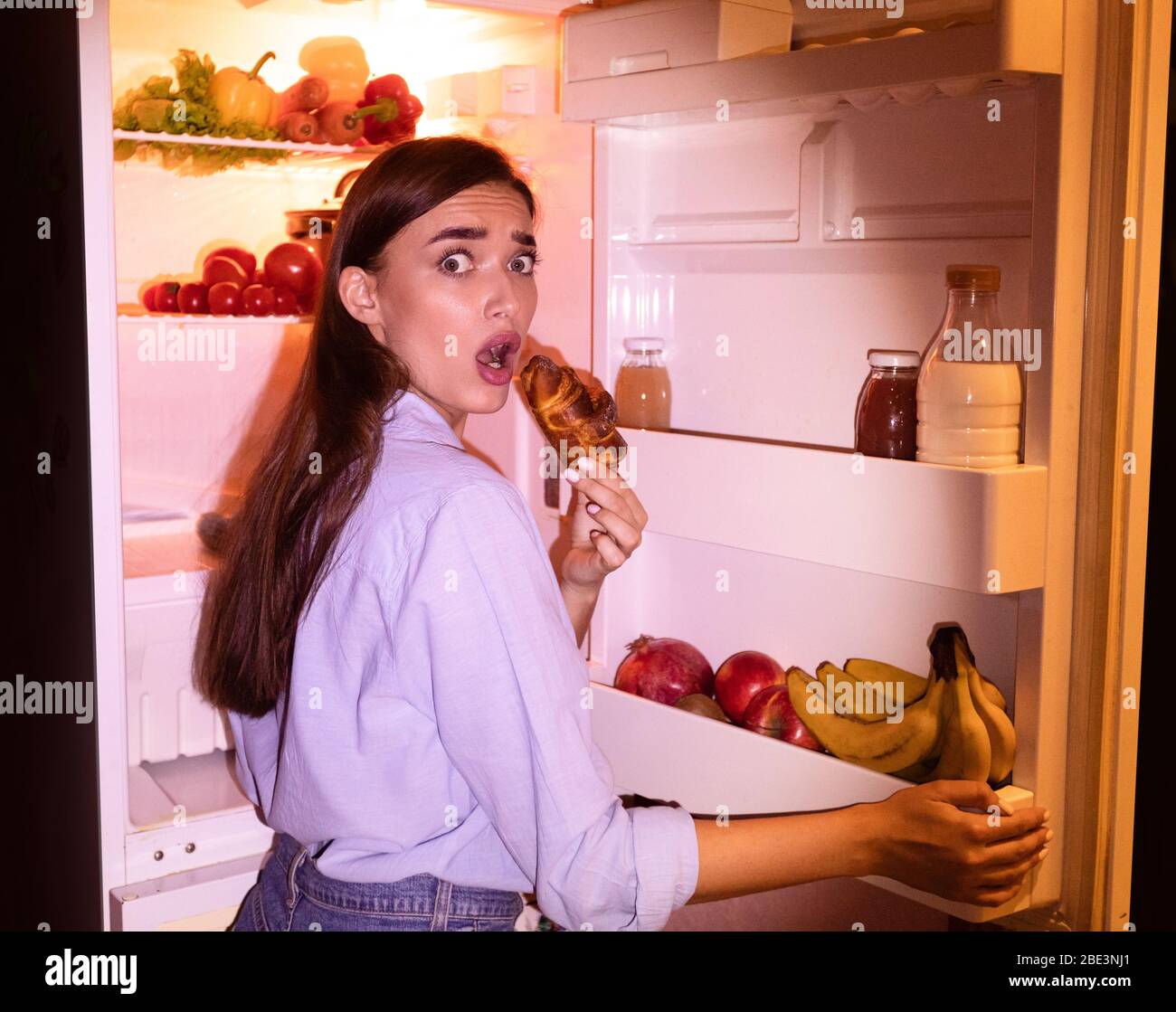 Young woman eating junk croissant near fridge in night Stock Photo