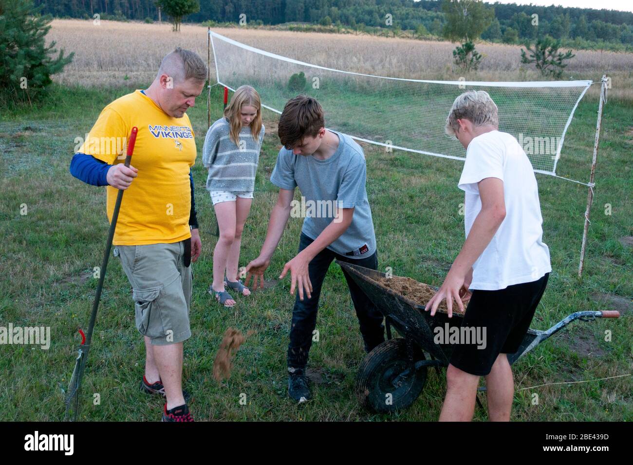 Father, sons and daughter with wheelbarrow of sand filling holes in badminton court. Zawady Gmina Rzeczyca Poland Stock Photo