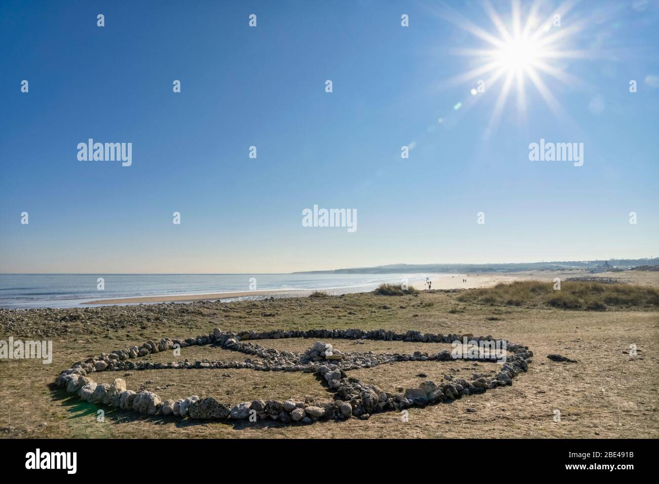 Symbolic rock formation of an X inside a circle made on the grass along the coast with people on the beach in the background Stock Photo