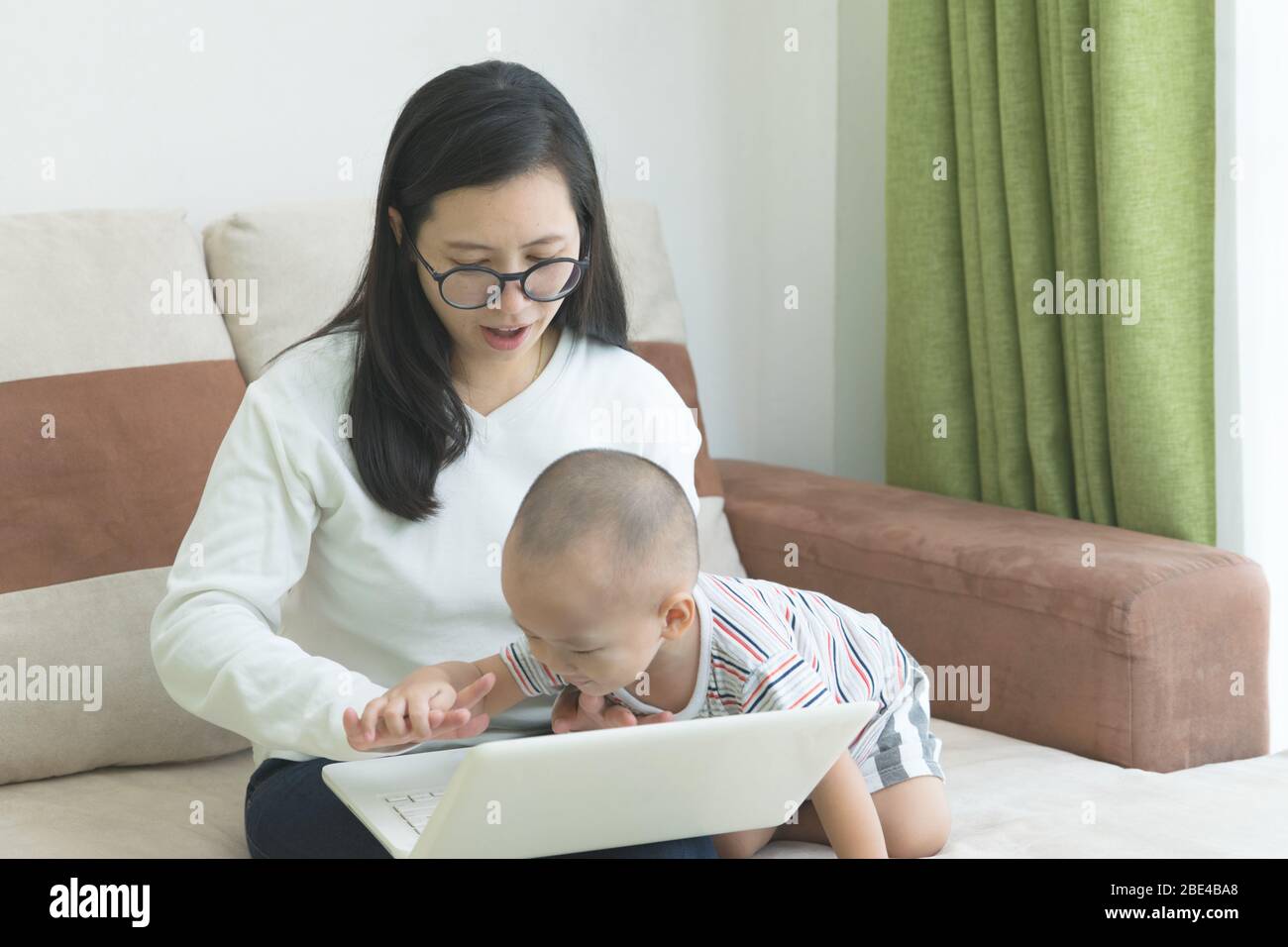 Young beautiful woman sitting on the sofa has disturbing by her kid while teleworking on her laptop . Work from home for social distancing concept . Stock Photo