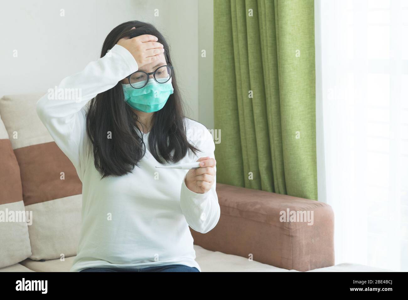 Sick young beautiful woman wearing green medical mask touch her head and check body temperature from thermometer in living room background. Coronaviru Stock Photo