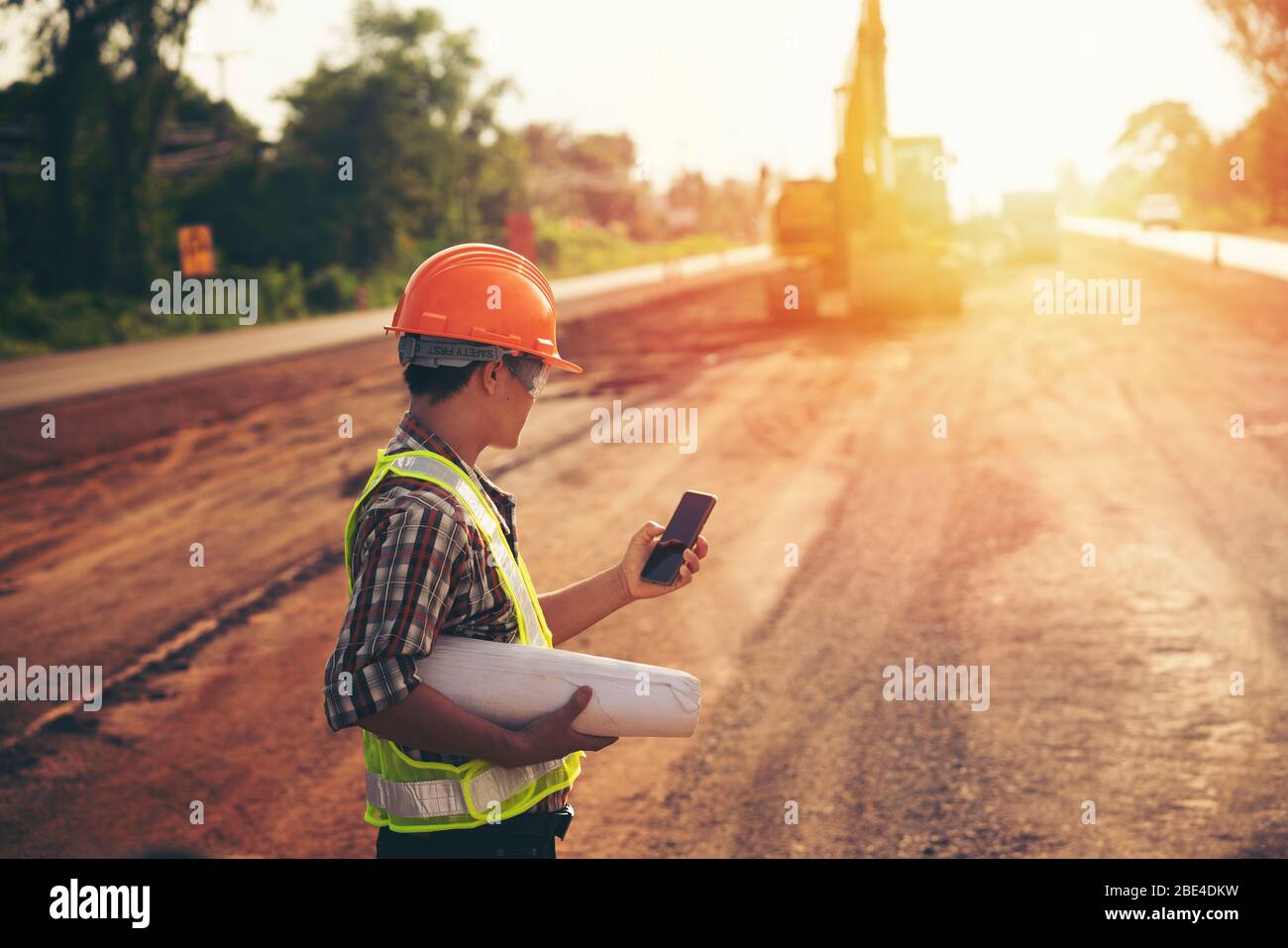 Engineer wear a hard hellmet and holding blueprint on road construction site with machinery,safety first concept Stock Photo
