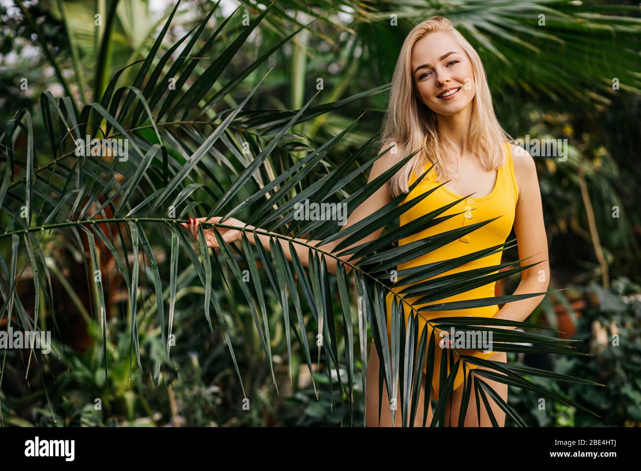 Young sexy woman with slim body, wearing yellow swimsuit standing among tropical trees. Horizontal shot of charming girl smiling posing in green leave Stock Photo