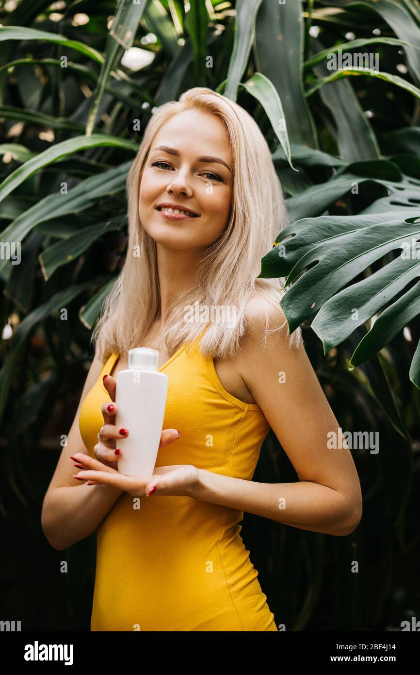 Gorgeous caucasian blonde woman with perfect clean skin, in yellow swimsuit holding cosmetic product, bottle of sunscreen lotion, standing among tropi Stock Photo