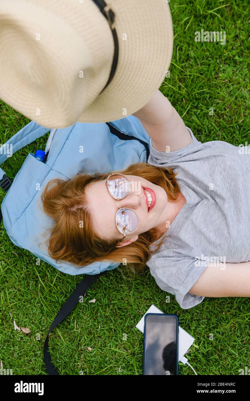 Happy young female traveller lay on backpack with smartphone, power bank and hat with palm trees reflection at her sunglasses, hold summer hat Stock Photo