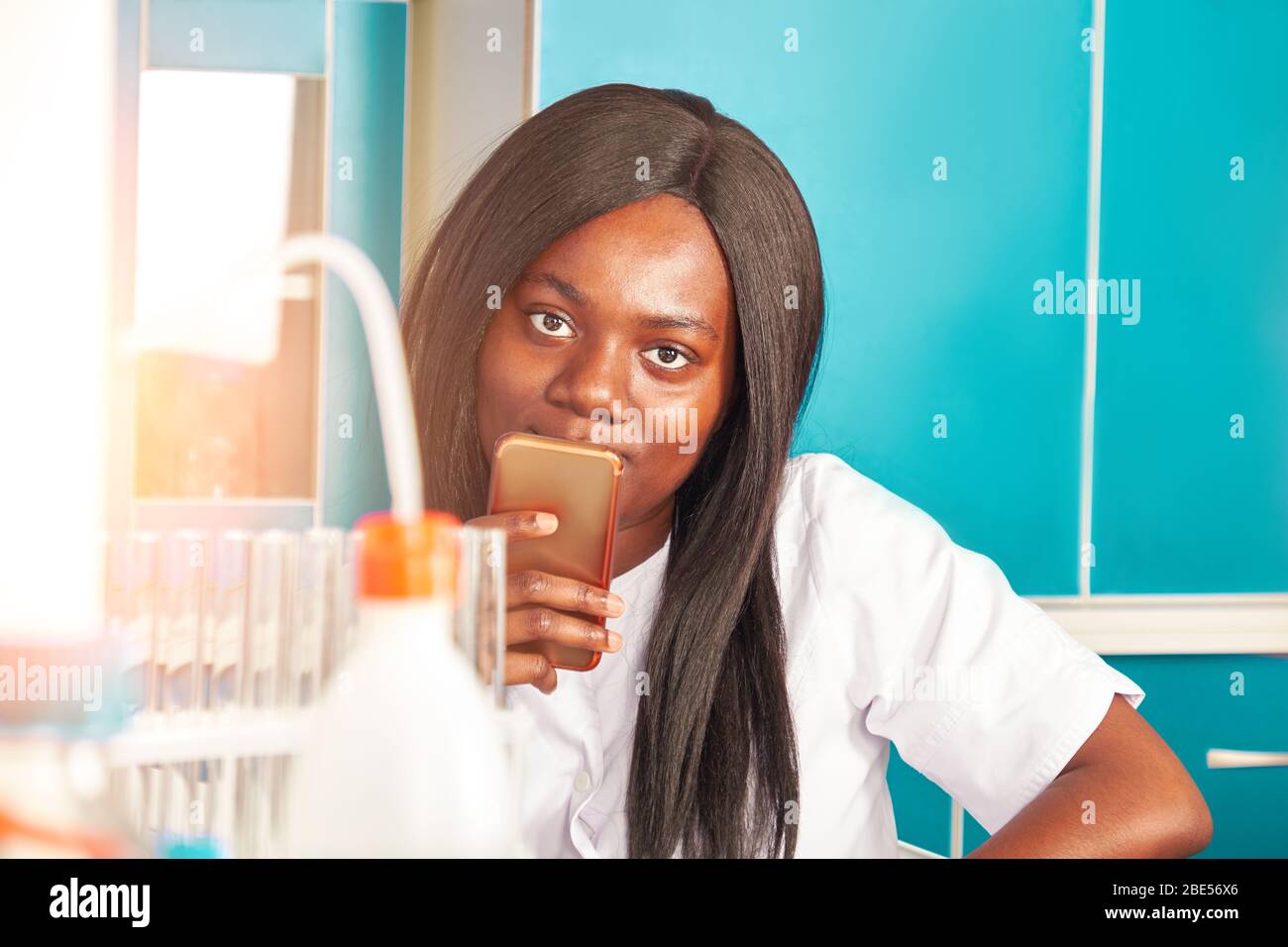 Young smiling African female scientist, graduate or medical student with mobile phone looks at the camera. The woman smiles while using digital device Stock Photo