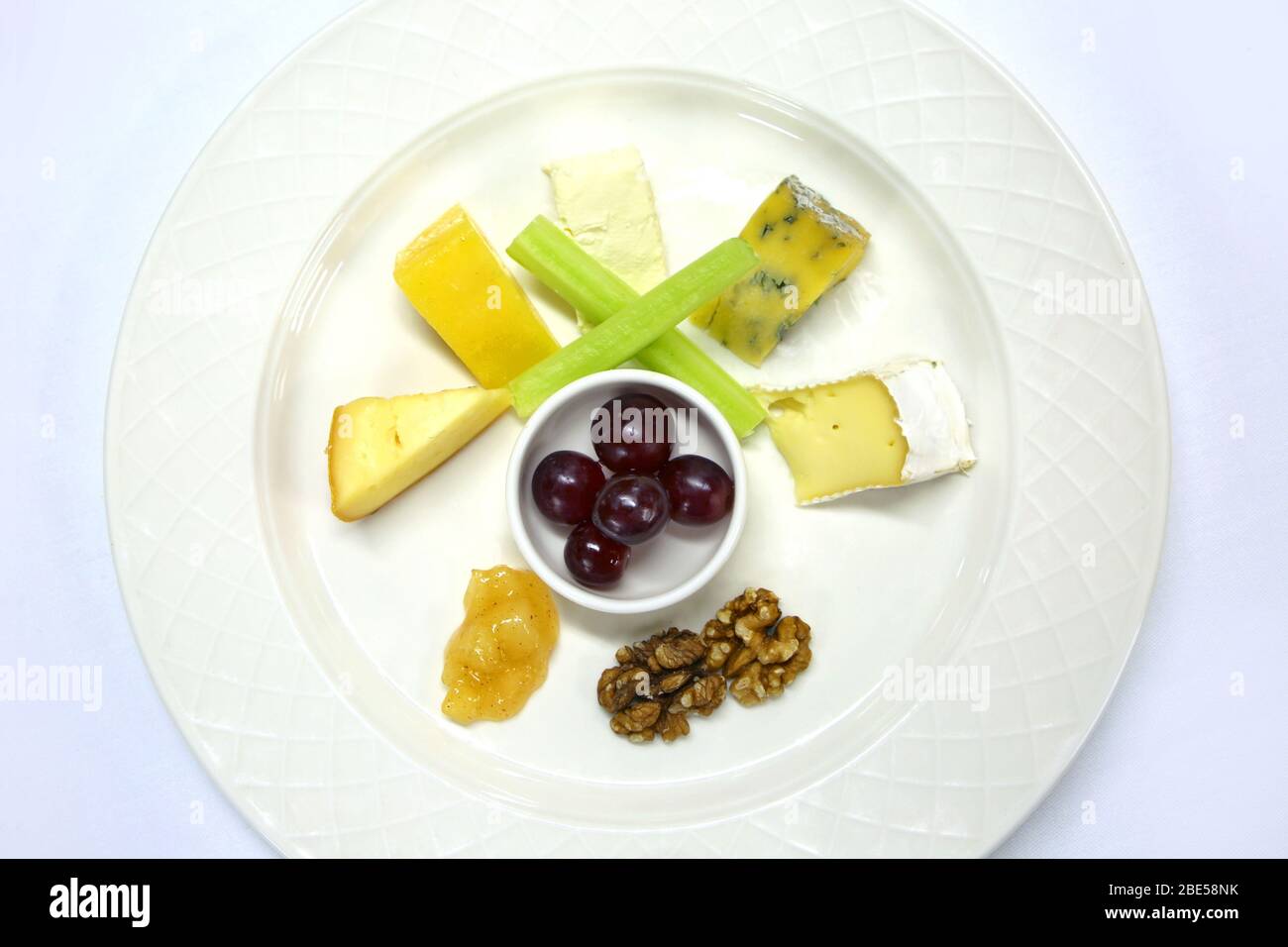 A selection of cheeses with grapes, chutney, celery & walnuts all prepared to serve as a cheese plate, on a white table cloth. Stock Photo