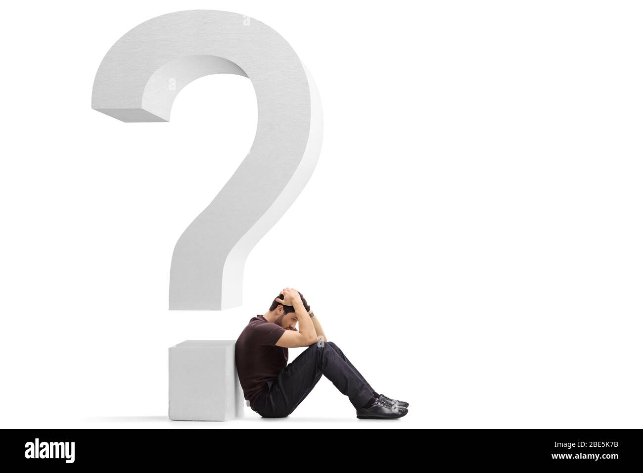 Sad young man sitting on the floor holding his head and leaning against a big question mark isolated on white background Stock Photo