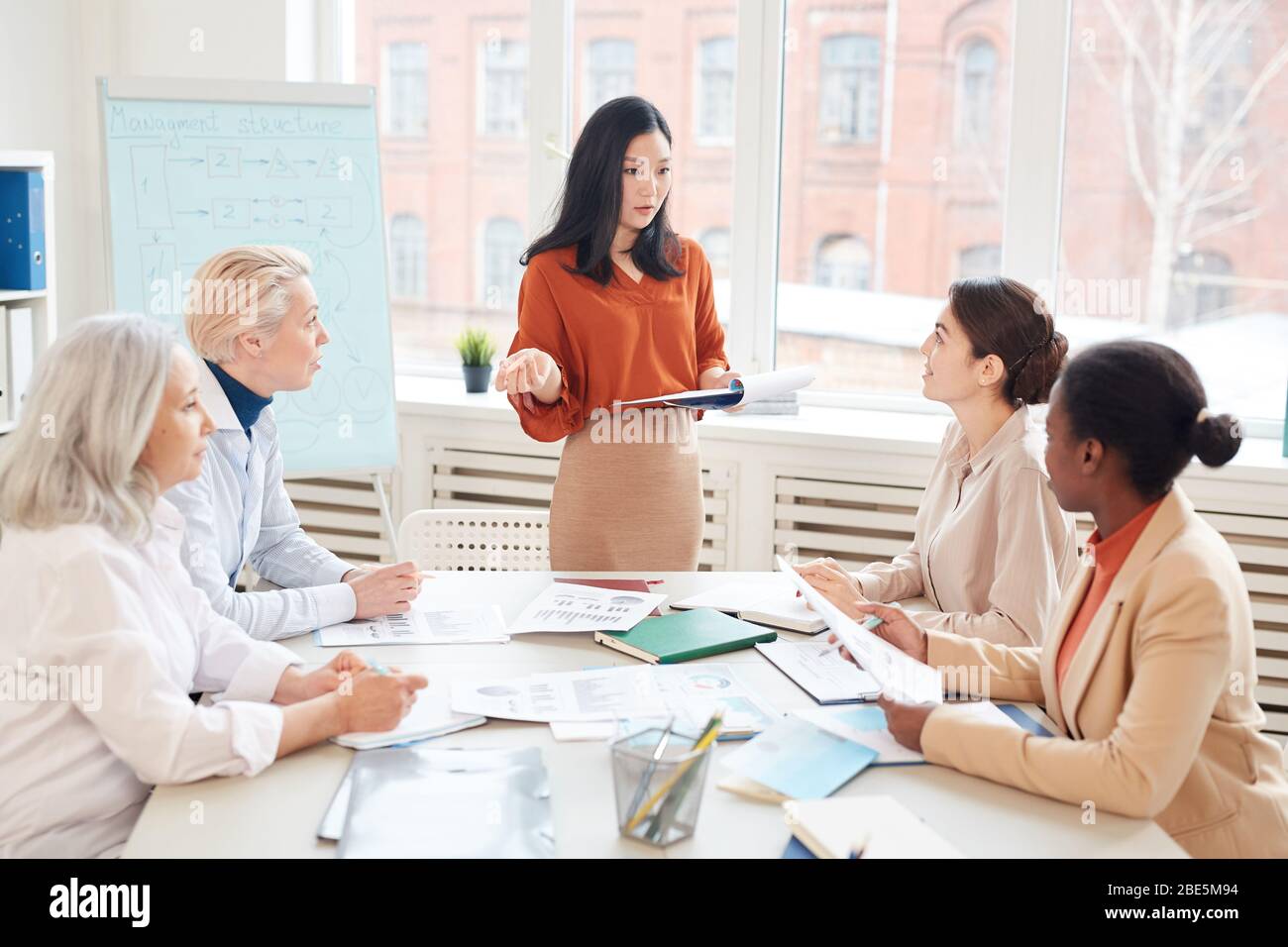 Portrait of successful Asian businesswoman presenting project plan to group of female colleagues while standing by whiteboard during meeting in confer Stock Photo