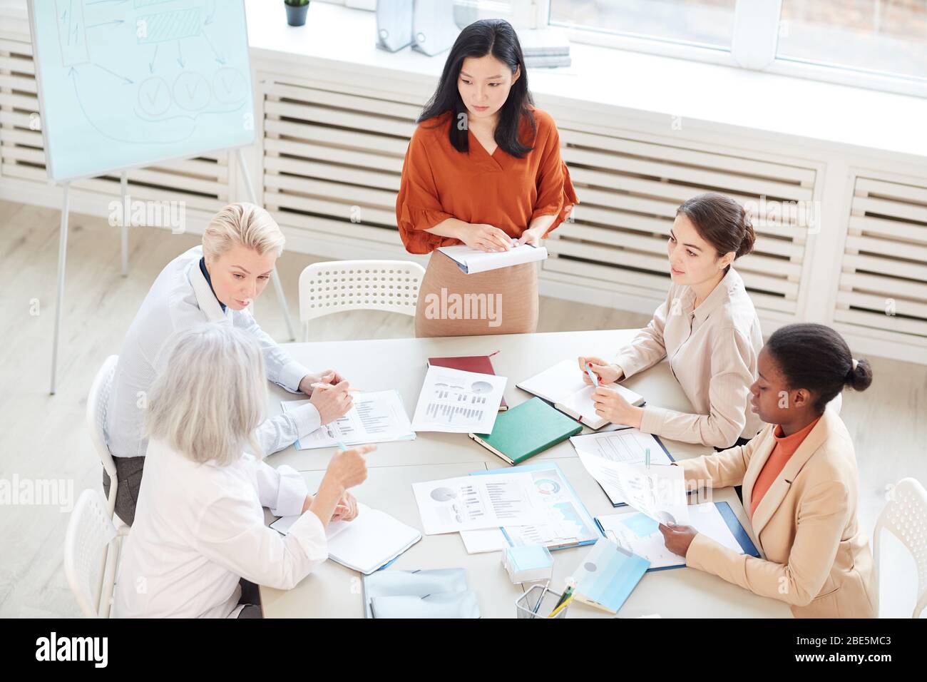 High angle view at successful Asian businesswoman presenting project plan to group of female colleagues while standing by whiteboard during meeting in Stock Photo