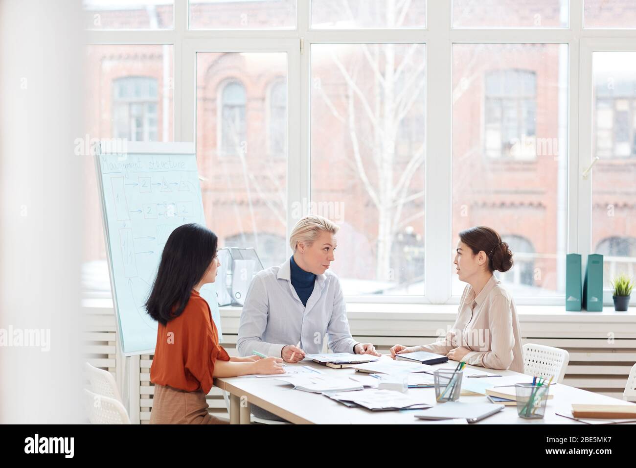Group of modern businesswomen discussing project while sitting at table against window during meeting in conference room, copy space Stock Photo