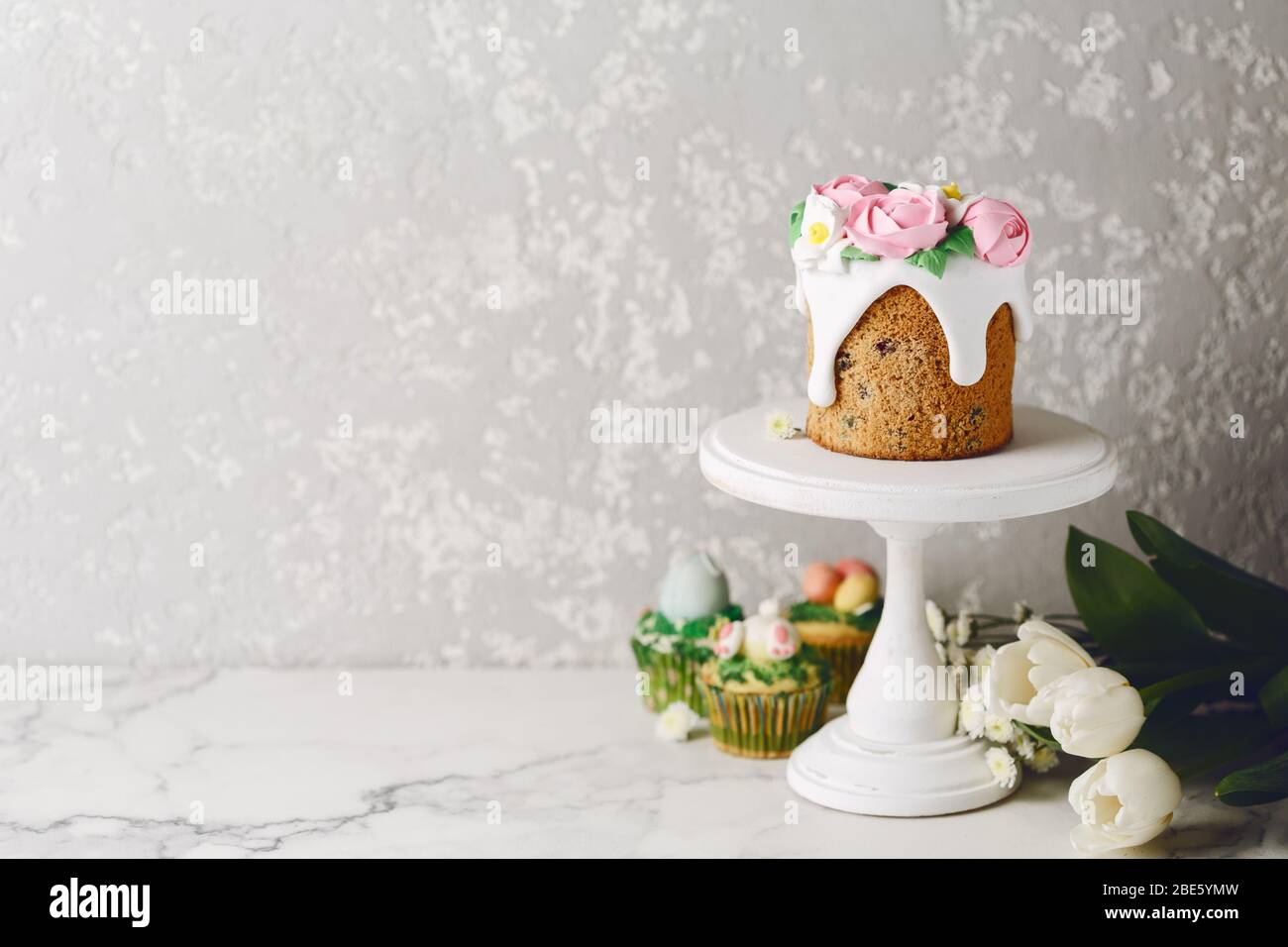 Easter cake with creamy roses on white stand. White flowers with holiday cupcakes. Cement wall. Copy space Stock Photo