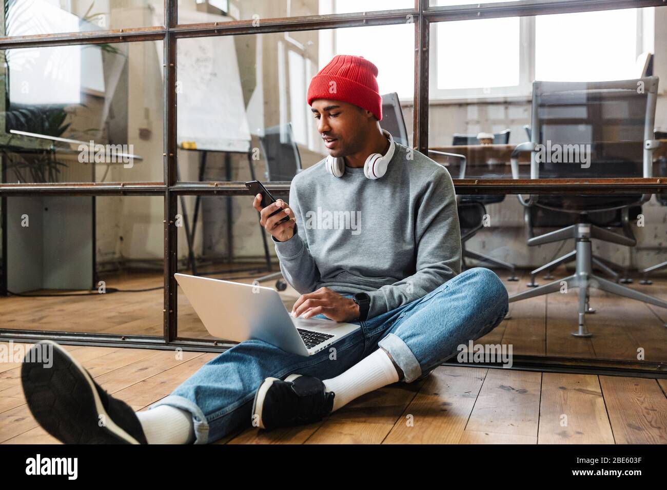 Image of a concentrated african young man sit indoors on floor using laptop computer and mobile phone. Stock Photo