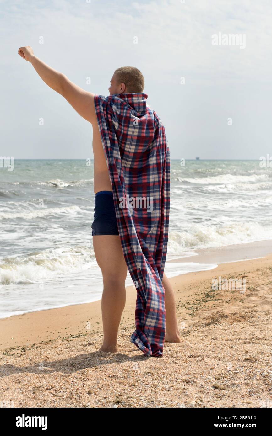 A funny fat man superhero with a plaid instead of a cloak stands on the sea beach. Stock Photo
