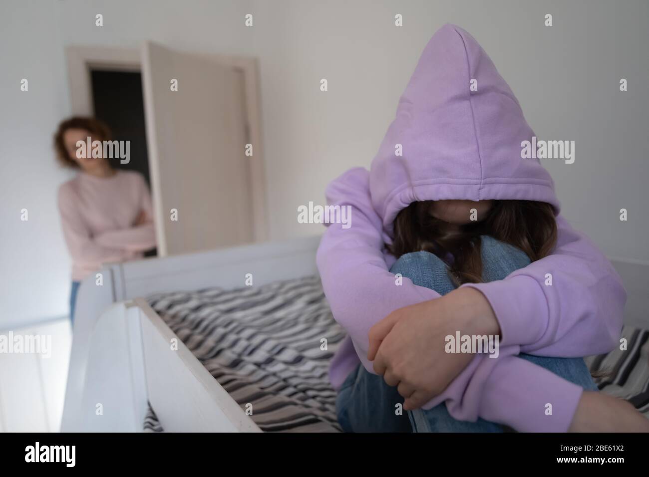 Depressed teen girl wearing hood sitting on bed ignoring mother Stock Photo