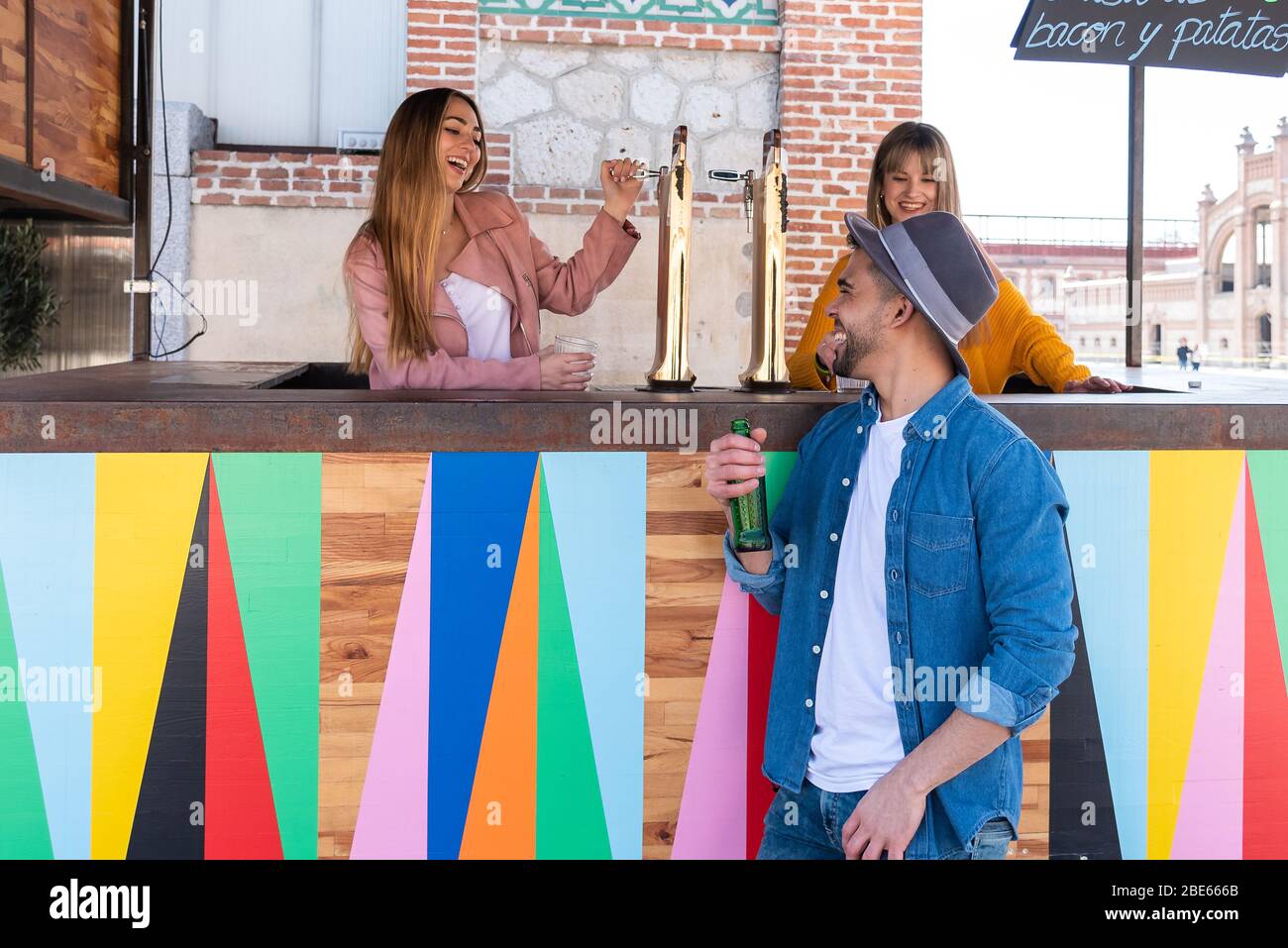 Two young blond-haired Caucasian women working at an outdoor bar toast and celebrate with a young Hispanic customer wearing a hat Stock Photo