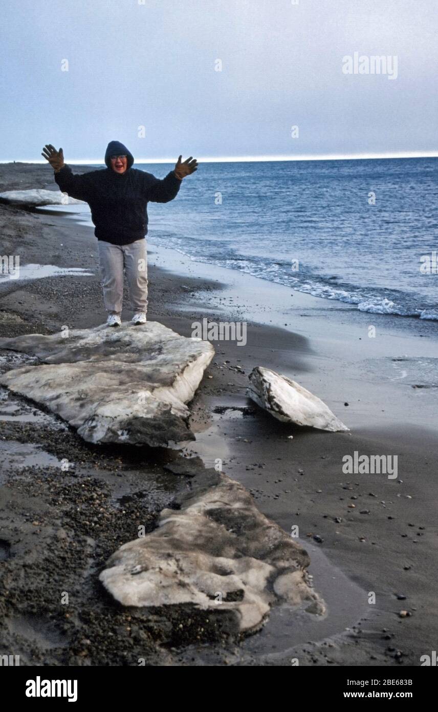Inuit woman stands on snow along the shore of the Arctic Ocean at midight on a summer evening with the a sliver of light in the horizon and diffuse li Stock Photo