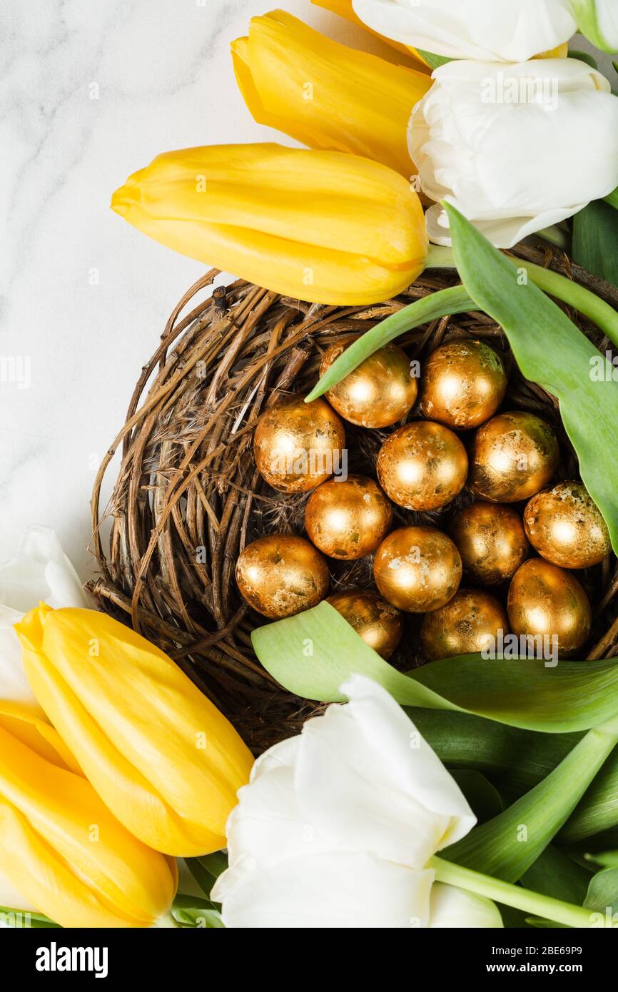 Beautiful bouquet of yellow and white tulips on marble table. Golden quail painted eggs in rustic nest. Happy Easter concept. Top view. Copy space Stock Photo