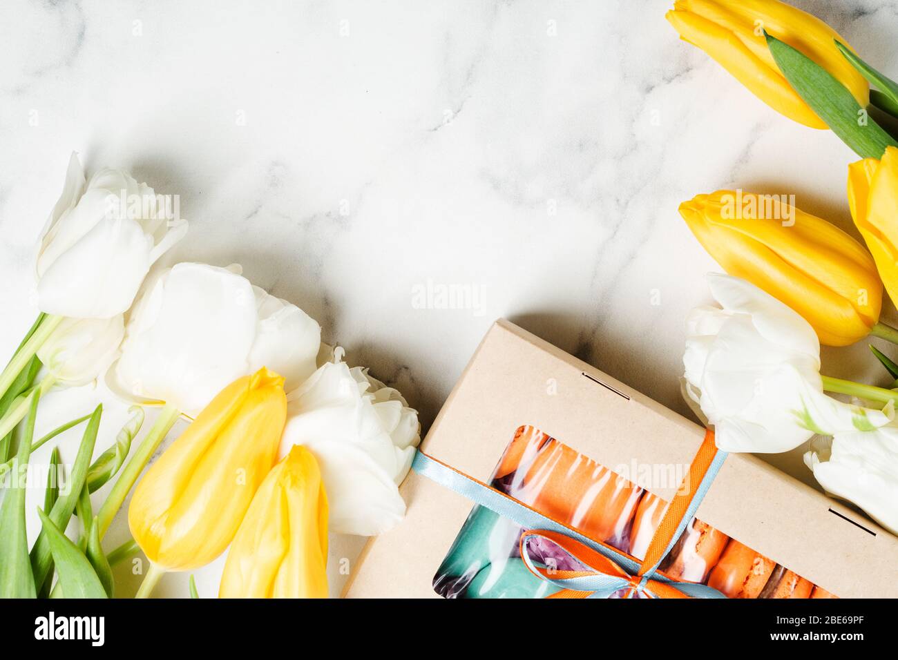 Box with orange and turquoise macaroons sweeties on marble table with bouquet of white and yellow tulips. Holiday concept Stock Photo