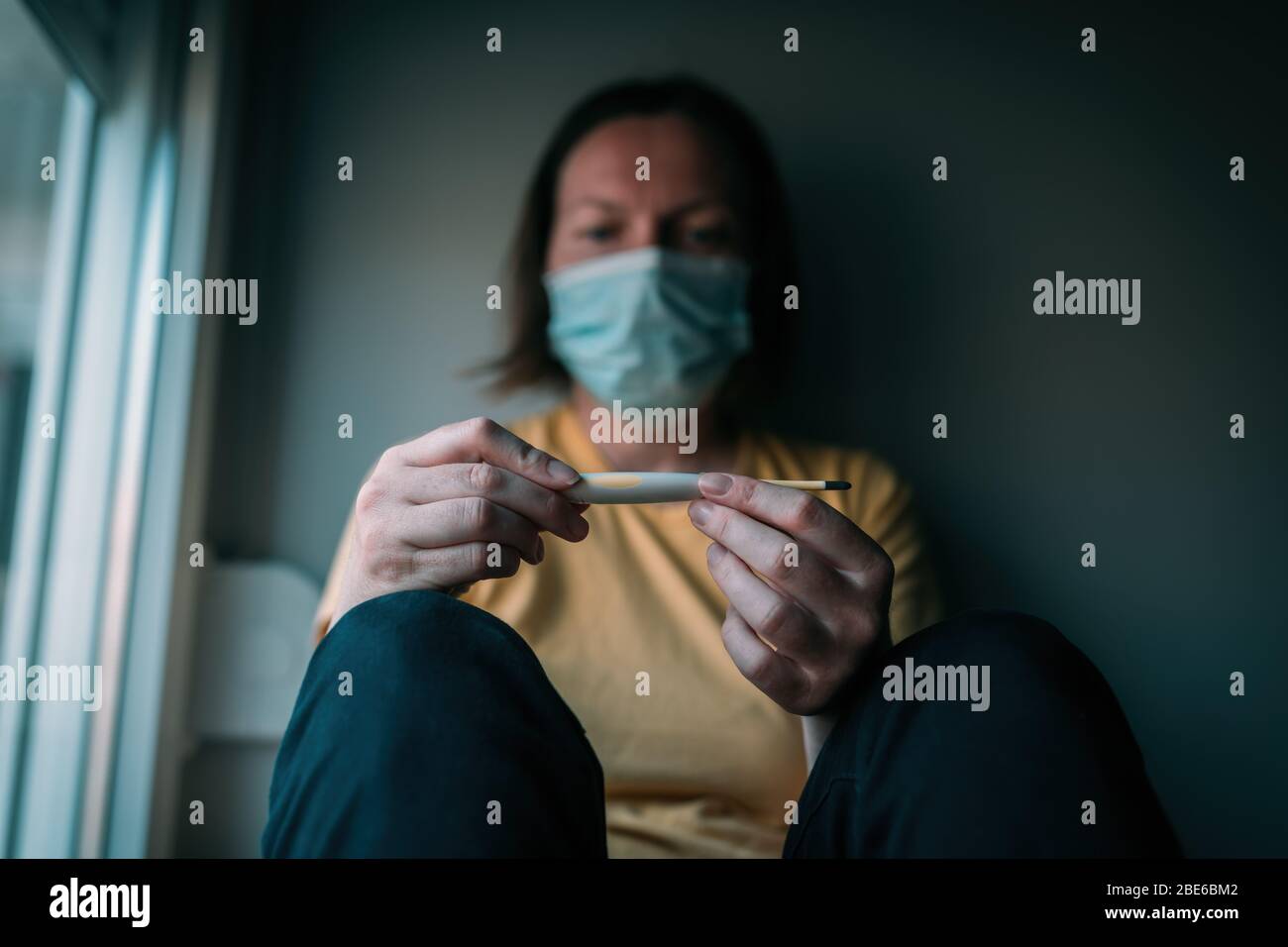 Woman in self-isolation measuring body temperature electronic digital thermometer. Female person with face protective respiratory mask, selective focu Stock Photo
