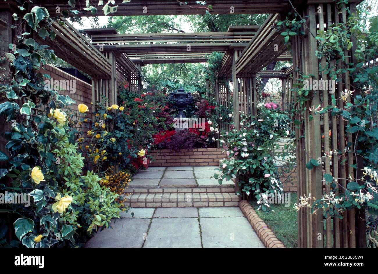 Formal paved  pergola with roses, honeysuckle and  ivy leading a raised bed with sculpture and massed azeleas. Stock Photo
