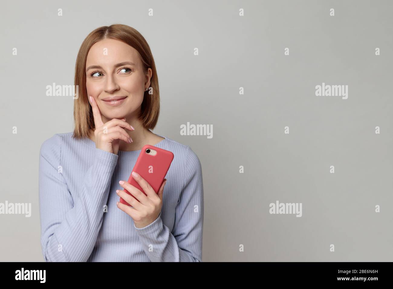 Studio portrait of pleasant cheerful lovely european woman looks up at blank copy space, holding smartphone, isolated over beige background. Positive Stock Photo
