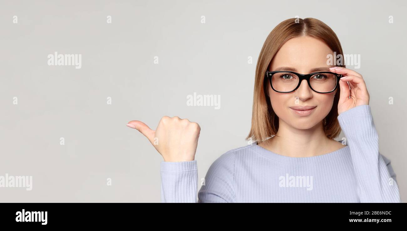 Close up portrait of successful European female with glasses, indicates with thumb at blank copy space, shows place for your advertisement, isolated Stock Photo