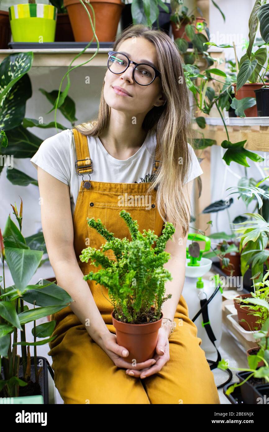 Woman gardener in glasses wearing overalls, holding a fern in plastic pot, potted houseplants on background. Home gardening, love of houseplants, free Stock Photo