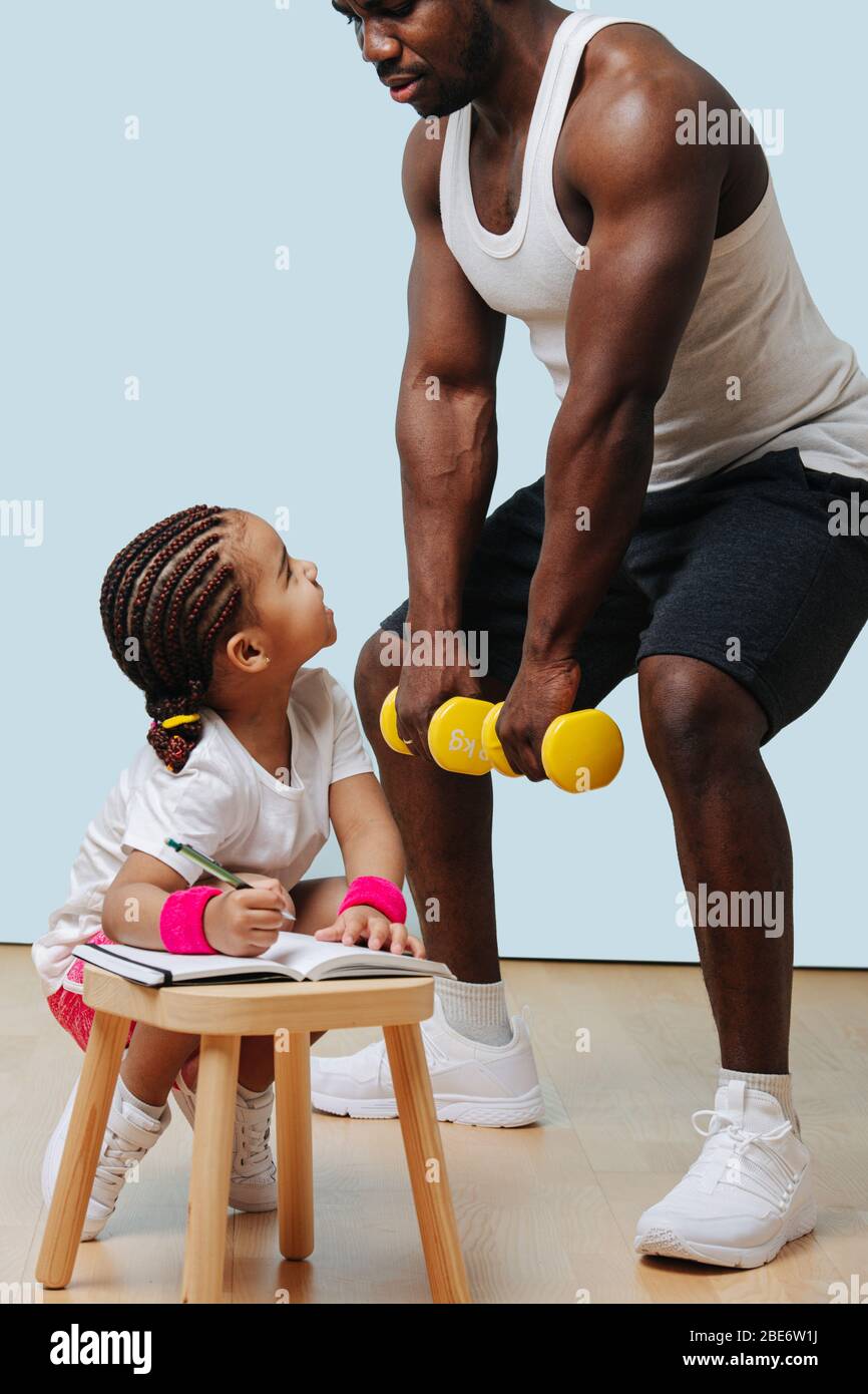 Black father doing squats, his daughter taking notes. Stock Photo
