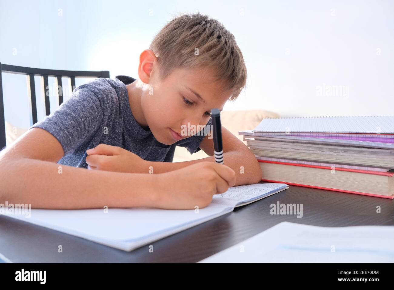 Child sitting among pile of books, textbooks, school exercise books on his desk and doing homework. Education, online distance learning for kids Stock Photo