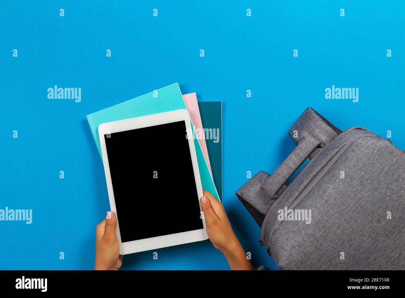 Kid hands with tablet computer, notebooks, books and gray backpack over light sky blue background Stock Photo