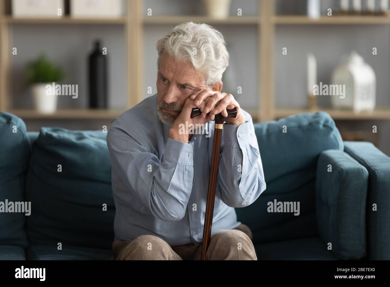 Tired upset mature man holding walking stick, sitting on couch Stock Photo