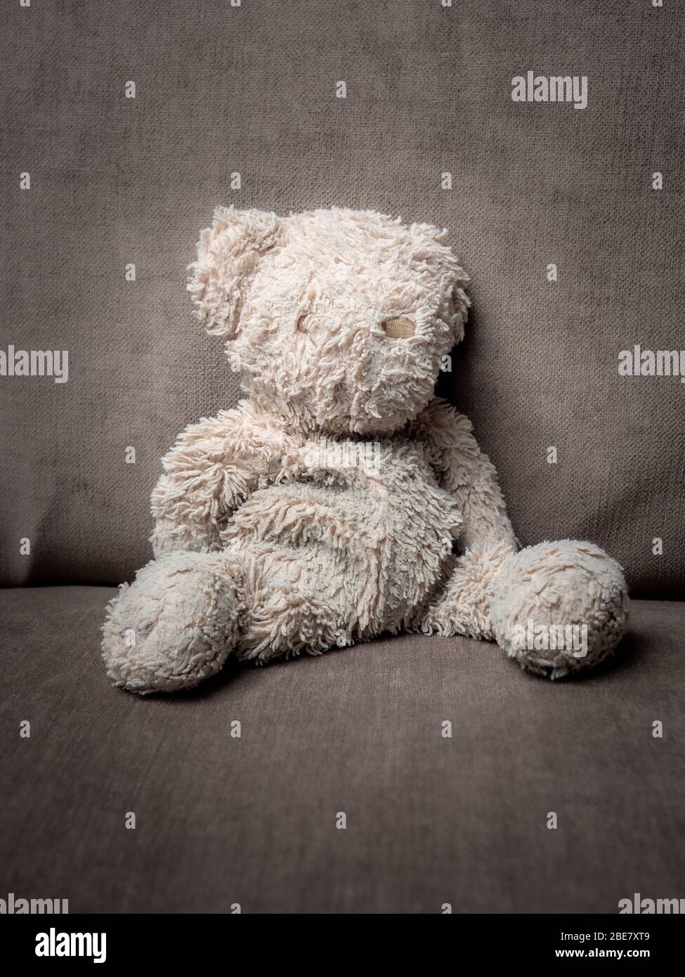 A tired, old teddy bear sitting, collapsed on a grey cushion Stock Photo