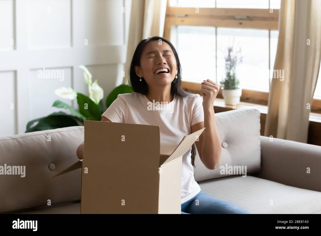 Asian woman sitting on sofa opens delivered parcel feels overjoyed Stock Photo