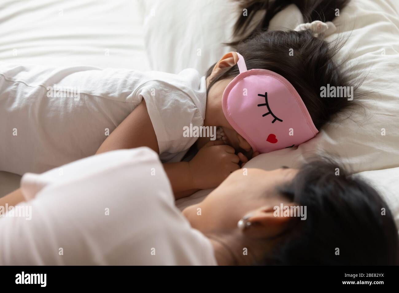 Small girl wears sleeping eyemask lying in bed with mother Stock Photo