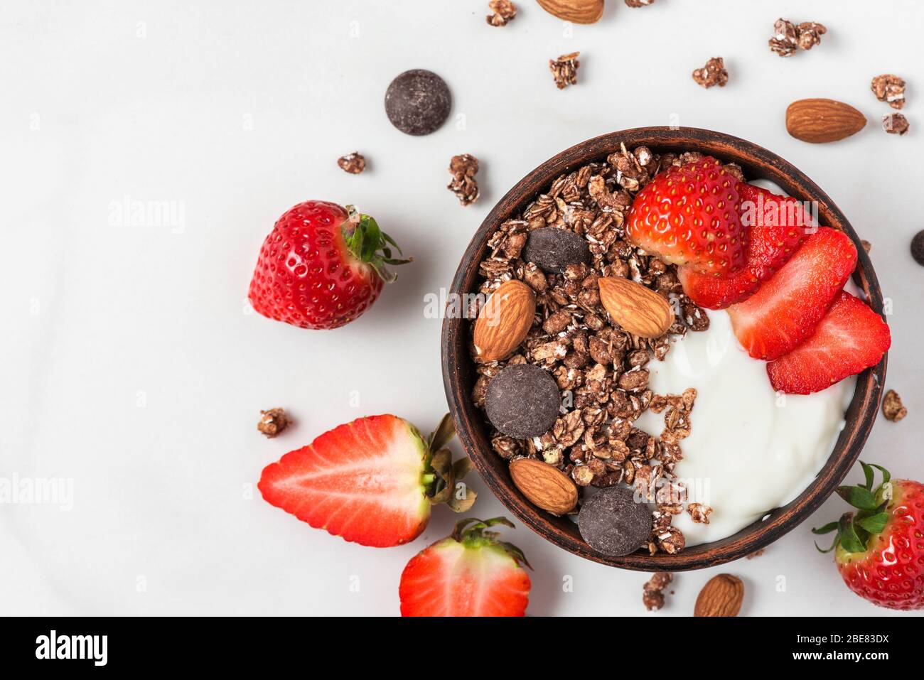 bowl of oat chocolate granola with yogurt, fresh strawberries and nuts on white marble background for healthy breakfast. top view Stock Photo