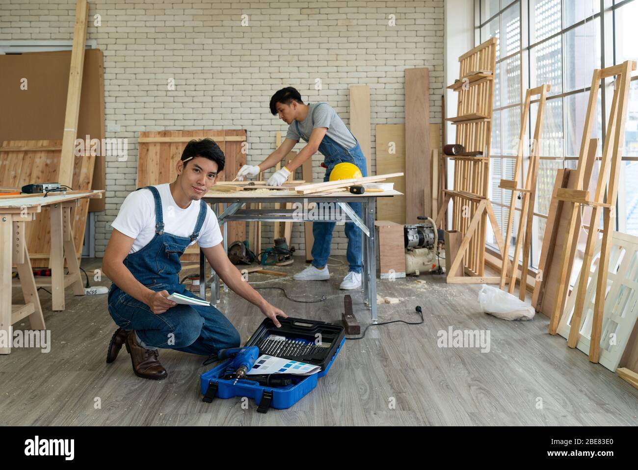 Young asian carpenter sat with the equipment confidently in his workshop. Another man using a pencil to draw drawings on a piece of wood. Stock Photo