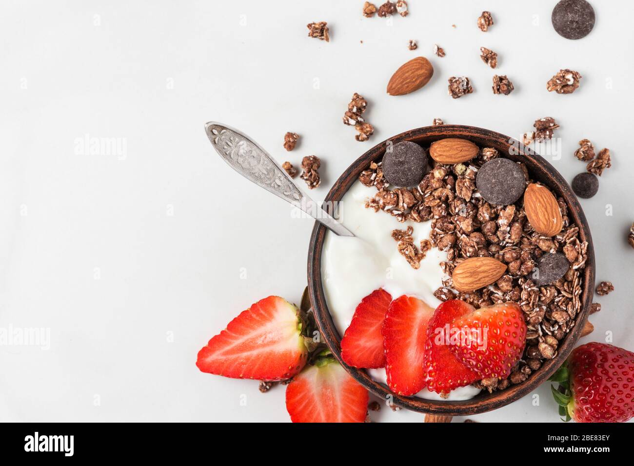 bowl of oat chocolate granola with yogurt, fresh strawberries and nuts with a spoon on white marble background for healthy breakfast. top view with co Stock Photo