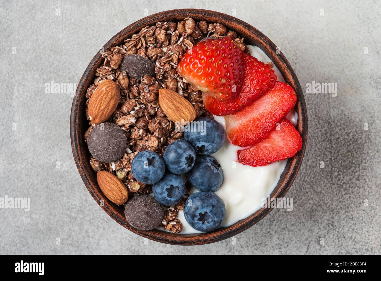 bowl of oat chocolate granola with yogurt, fresh blueberries, strawberries and nuts on gray background for healthy breakfast. top view Stock Photo