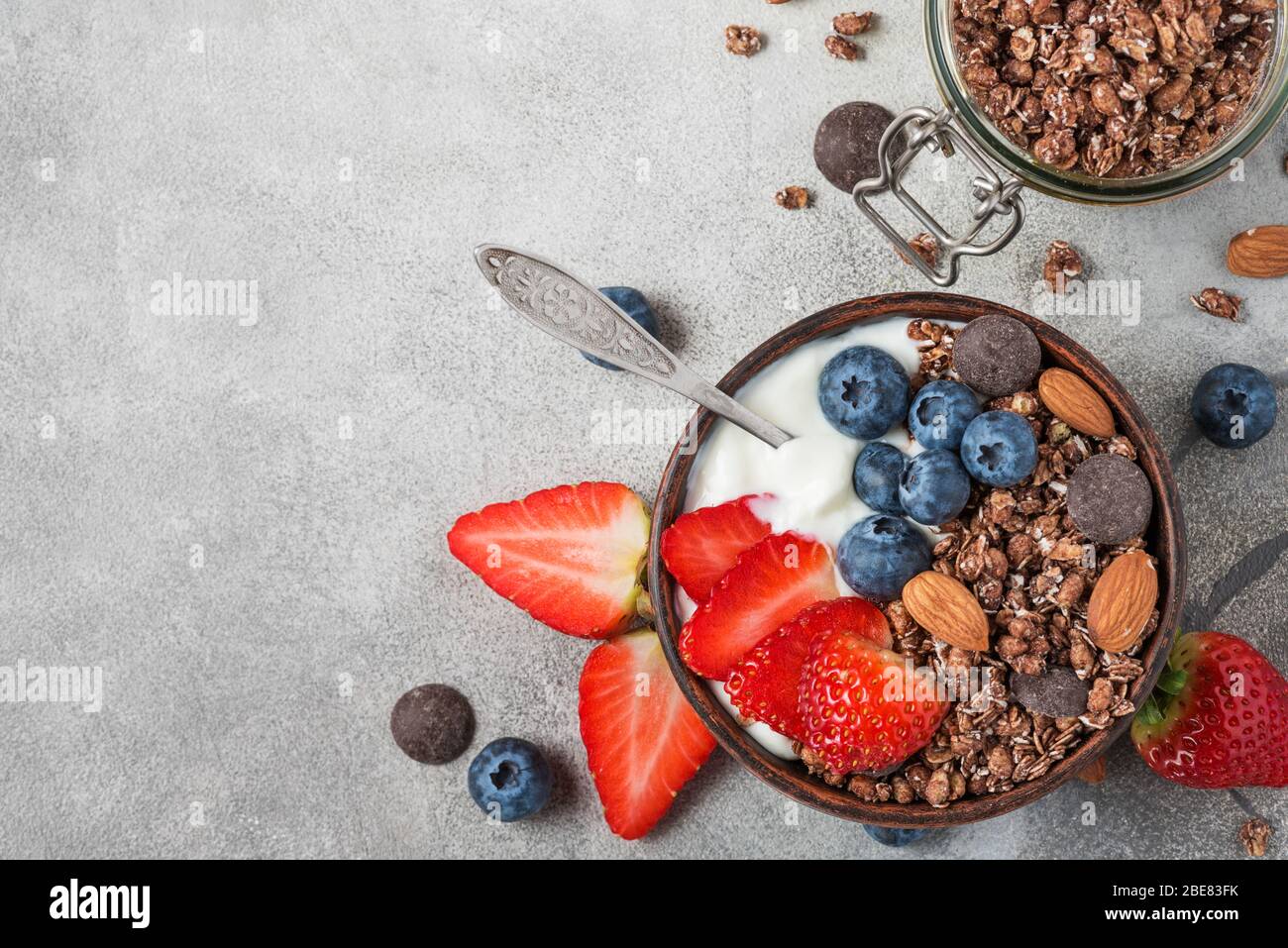 bowl of oat chocolate granola with yogurt, fresh blueberries, strawberries and nuts with a spoon on gray background for healthy breakfast. top view wi Stock Photo